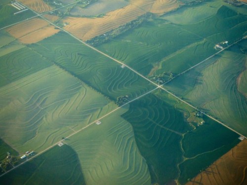 Aerial view of patchwork farmland with wavy patterns in green and brown fields, intersected by roads.