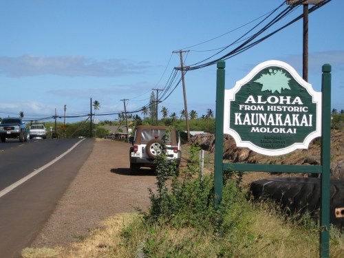 Sign reading "Aloha from Historic Kaunakakai, Molokai" beside a road with parked vehicles and palm trees in the background.