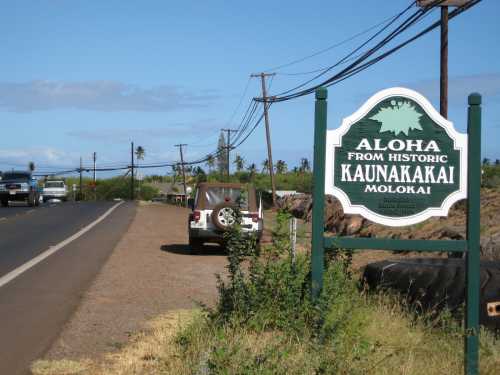 Sign reading "Aloha from Historic Kaunakakai, Molokai" beside a road with parked vehicles and palm trees in the background.