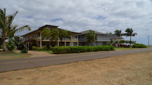 A large, two-story house with palm trees and a grassy area, set along a road near the beach under a cloudy sky.