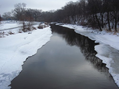 A calm river reflects trees, bordered by snow and ice, under a cloudy sky in a winter landscape.