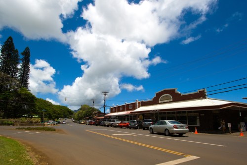 A sunny street scene with shops, cars parked, and fluffy clouds in a blue sky. Lush greenery in the background.
