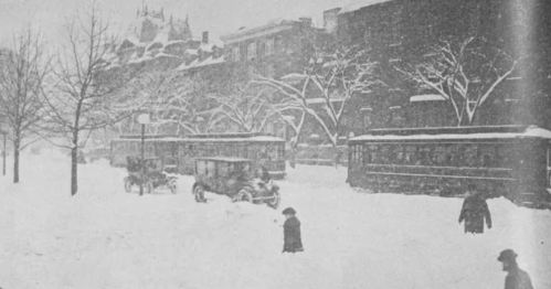 Historic snowy street scene with vintage cars and streetcars, trees bare, and people walking through deep snow.