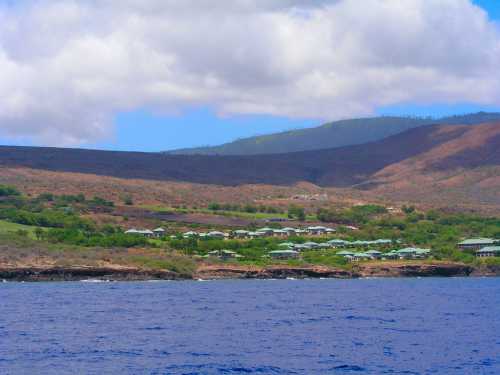 Coastal view of a green hillside with buildings, under a partly cloudy sky and blue ocean in the foreground.