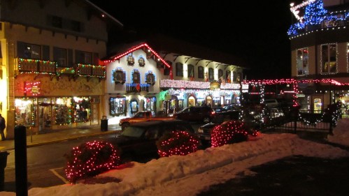 A festive street scene at night, adorned with colorful holiday lights and decorations, with snow on the ground.