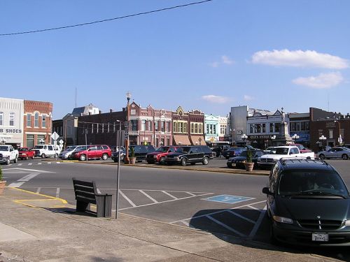 A small town square with historic buildings, parked cars, and a clear blue sky. Benches line the street.