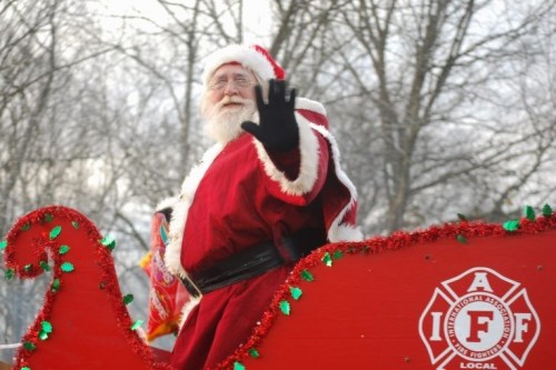 Santa Claus waves from a festive sleigh decorated with red and green, surrounded by winter trees.