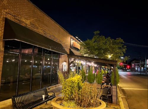 A cozy outdoor dining area at night, featuring benches, plants, and warm lighting near a brick building.