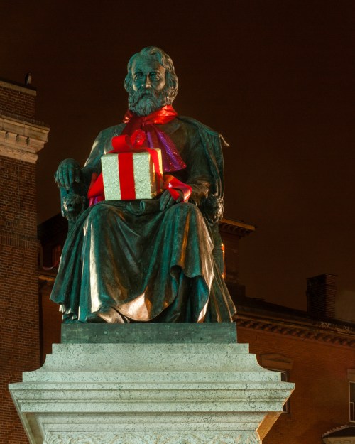 A statue of a seated figure wearing a red bow, holding a gift box, illuminated at night.