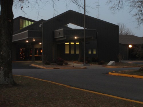 A modern, dark-colored building with large windows, surrounded by trees and a paved driveway, at dusk.