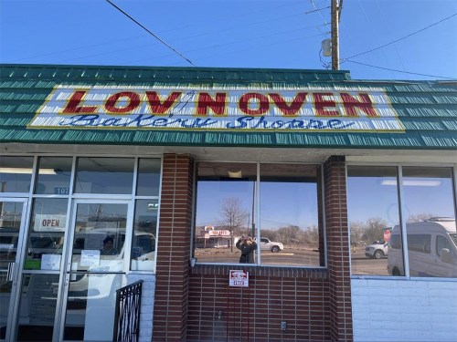 A storefront with a green roof and a sign that reads "LOV NOVER" above large windows, reflecting the surroundings.