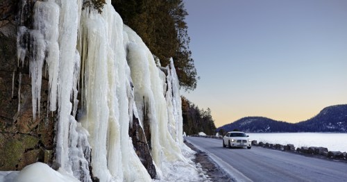 A car drives along a snowy road beside large icicles hanging from a rocky cliff near a lake at sunset.