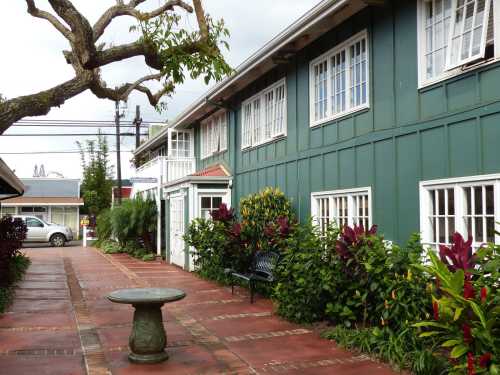 A green building with white windows, surrounded by colorful plants and a stone table in a paved courtyard.