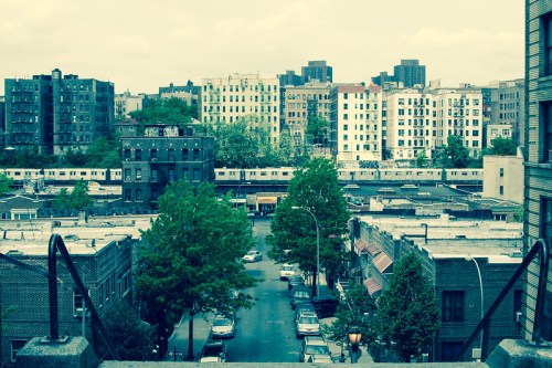 Urban landscape featuring buildings, trees, and a street with parked cars, under a cloudy sky.
