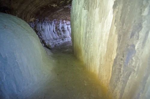 A narrow ice cave with glistening icy walls and a smooth, frozen floor illuminated by soft light.