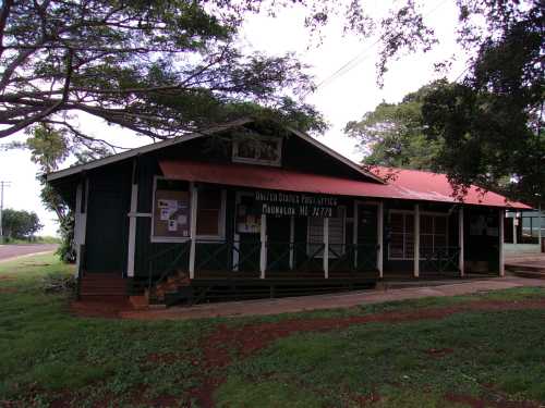 A green wooden building with a red roof, labeled "United States Post Office," surrounded by trees and grass.