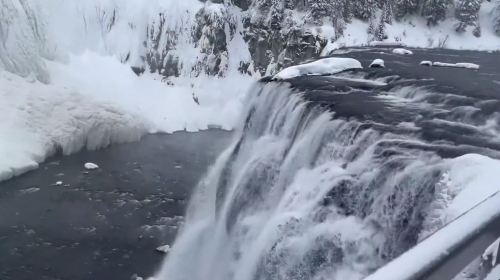 A snowy waterfall cascading into a dark river, surrounded by icy cliffs and a winter landscape.