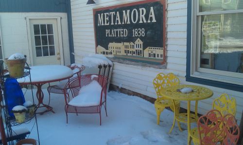 A snowy outdoor patio with colorful chairs and a sign reading "Metamora Platted 1838" on a white wooden wall.