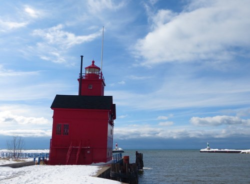 A red lighthouse stands by the water under a cloudy sky, with a snowy shoreline and distant piers visible.