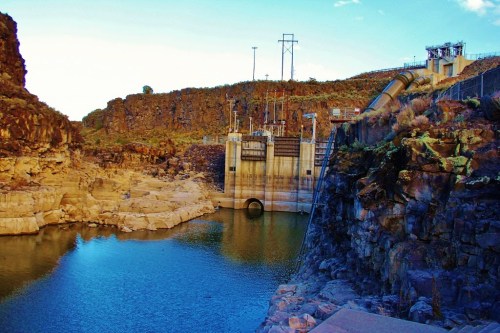 A dam structure beside a calm body of water, surrounded by rocky terrain and power lines against a clear blue sky.
