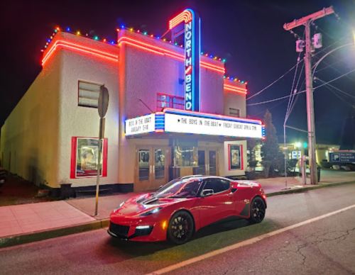 A red sports car parked in front of the brightly lit North Bend theater at night. Neon lights illuminate the building.