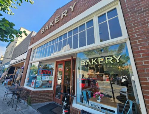 A charming bakery storefront with large windows, displaying "BAKERY" and "OPEN" signs, surrounded by outdoor seating.