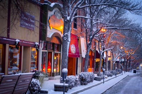 Snow-covered street lined with charming shops, glowing warm lights, and frosty trees in a winter setting.
