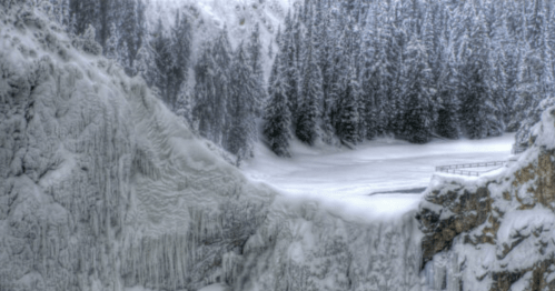 A snowy landscape featuring icy cliffs and a frozen lake, surrounded by tall evergreen trees.
