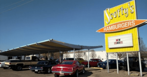 A retro diner named Scotty's Hamburgers with a bright sign, featuring classic cars parked outside under a canopy.
