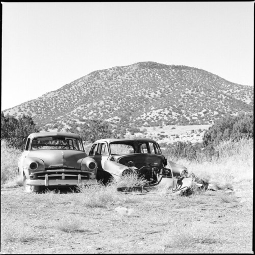 Two vintage cars sit abandoned in a grassy field, with a mountainous landscape in the background.