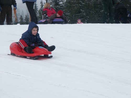 A child in a blue jacket sleds down a snowy hill, surrounded by other people enjoying winter activities.