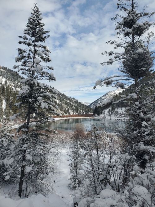 Snow-covered trees frame a serene lake surrounded by mountains under a cloudy sky.