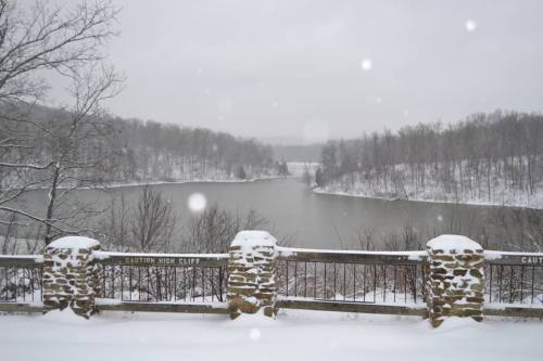 A snowy landscape featuring a river surrounded by trees, with a stone railing in the foreground. Snowflakes are falling.