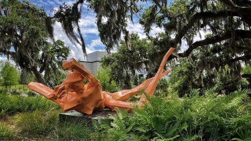 A vibrant orange sculpture of a reclining figure surrounded by lush greenery and hanging moss.