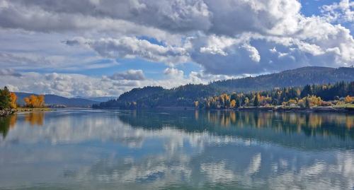 A serene lake reflects clouds and colorful autumn trees under a bright blue sky.