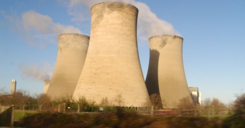 Three large cooling towers emitting steam against a clear blue sky, surrounded by greenery.
