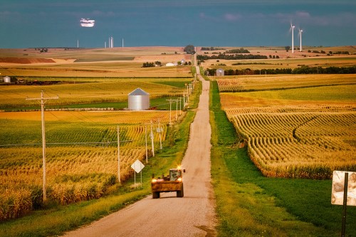 A rural road stretches through golden fields, with a tractor in the foreground and wind turbines in the distance.