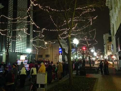 A festive street scene at night, lined with trees adorned with lights and people enjoying the holiday atmosphere.