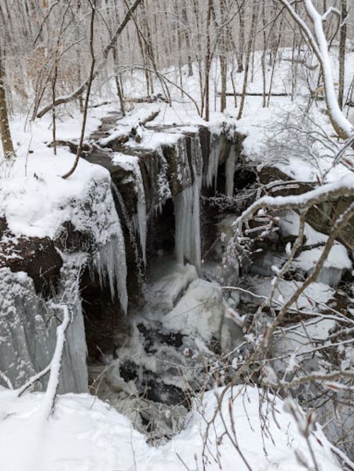 A snowy landscape featuring a frozen waterfall with icicles, surrounded by bare trees and a flowing stream below.