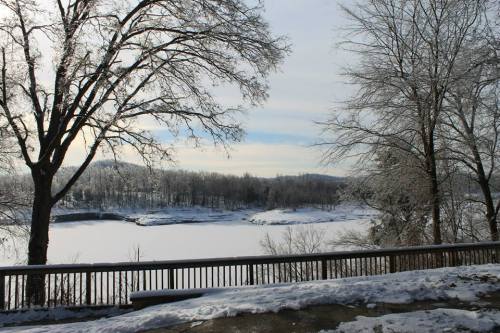 A snowy landscape with bare trees overlooking a frozen lake under a cloudy sky.