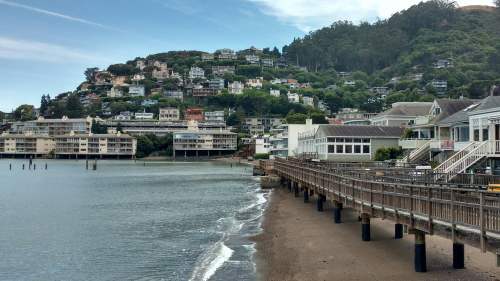 Scenic waterfront view with a wooden pier, sandy shore, and hillside homes in a coastal town.