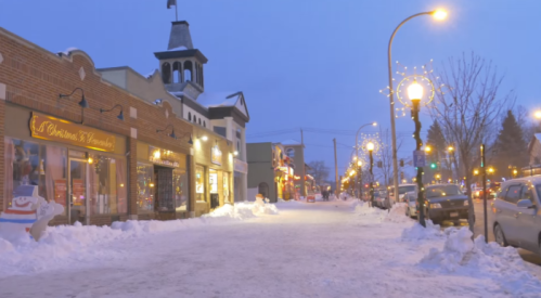 Snow-covered street lined with shops and festive lights, illuminated by street lamps at dusk.