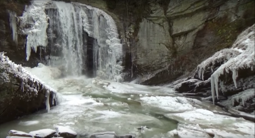 A frozen waterfall surrounded by icy rocks and a flowing river, creating a serene winter landscape.