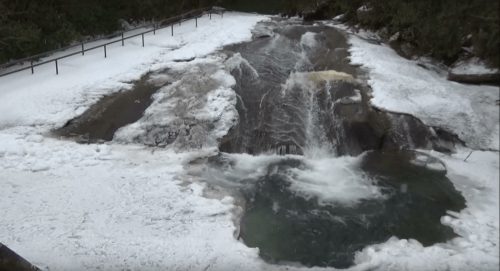 A frozen river with flowing water, surrounded by snow and rocky banks, under a forested area.