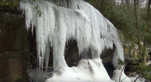 Icicles hang from a rocky cliff, with snow and ice covering the ground below in a serene winter landscape.