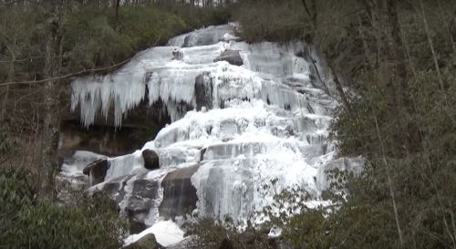 A frozen waterfall cascading down rocky cliffs, surrounded by trees and winter scenery.
