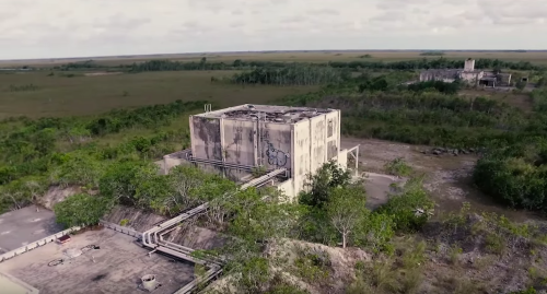 Aerial view of an abandoned industrial building surrounded by overgrown vegetation and open fields.