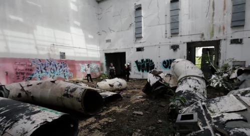 Abandoned industrial space with graffiti, scattered debris, and overgrown plants, featuring two people exploring.