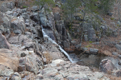 A rocky landscape featuring a small waterfall cascading into a pool, surrounded by sparse trees and dry foliage.