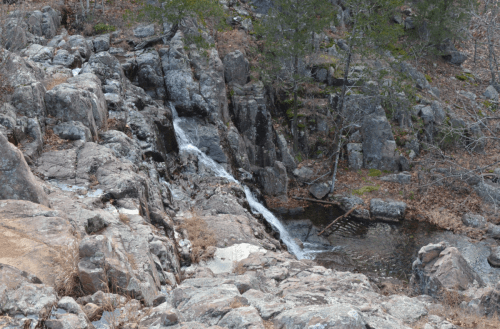 A rocky landscape featuring a small waterfall cascading into a pool, surrounded by sparse trees and dry foliage.